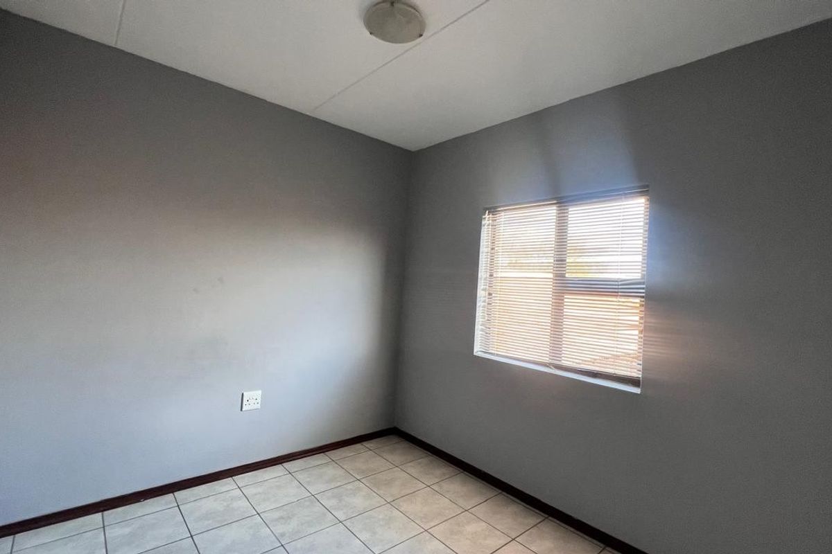 A clean, empty bedroom featuring neutral grey walls, tiled flooring, and a window fitted with venetian blinds.