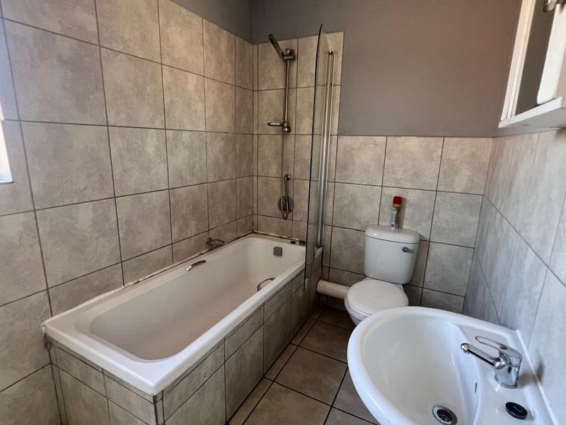 A functional bathroom featuring a bathtub with a shower attachment, glass screen, pedestal sink, and toilet, finished with neutral wall and floor tiling.