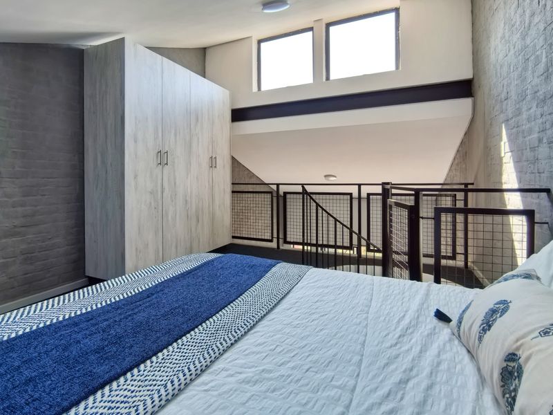 A modern loft-style bedroom featuring exposed brick walls, built-in cabinetry, and a metal staircase railing overlooking the lower level.