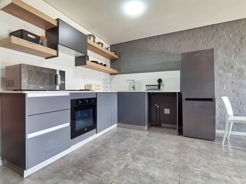 Modern kitchen featuring dark grey cabinetry, floating timber shelves, exposed brick feature wall, built-in oven, and sleek tiled flooring.