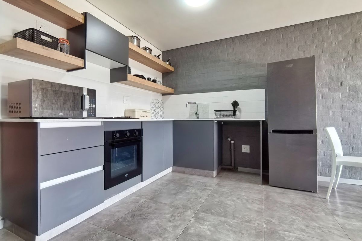 Modern kitchen featuring dark grey cabinetry, floating timber shelves, exposed brick feature wall, built-in oven, and sleek tiled flooring.