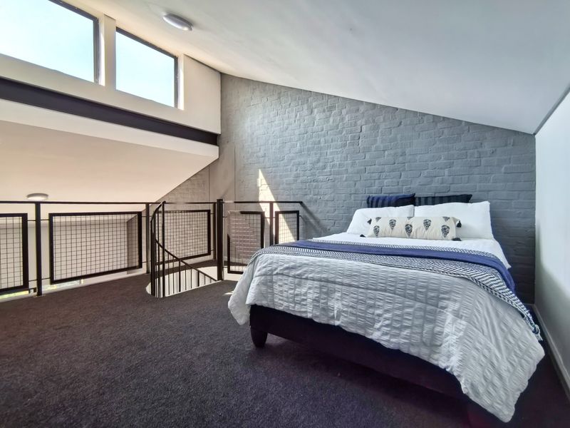 A spacious loft-style bedroom featuring an exposed grey brick wall, metal spiral staircase with mesh balustrade, and high clerestory windows providing natural light.