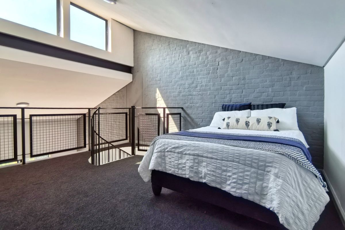 A spacious loft-style bedroom featuring an exposed grey brick wall, metal spiral staircase with mesh balustrade, and high clerestory windows providing natural light.