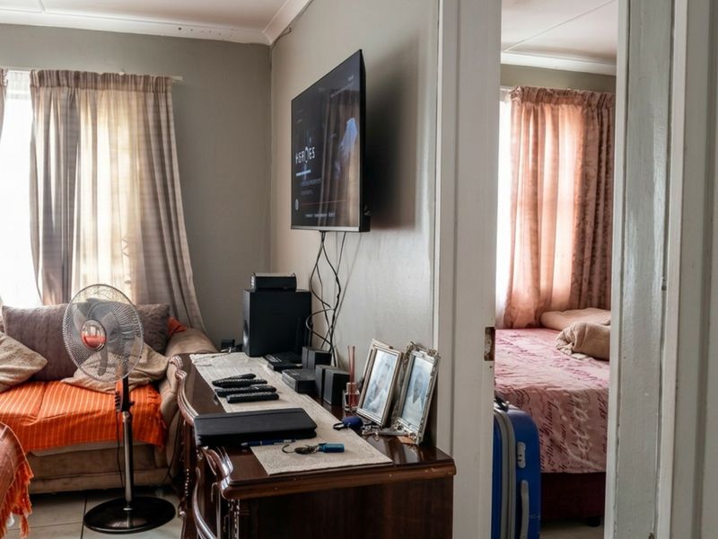 A living room area featuring tiled flooring, coved ceilings, and a wall-mounted television, with an adjacent view into a bedroom.