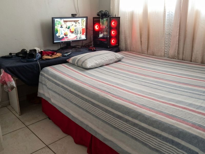 A compact bedroom featuring tiled flooring and window curtains, currently utilized as a combined sleeping and workspace area.