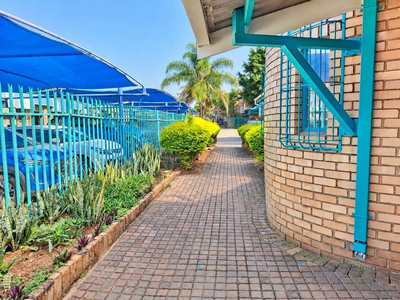A paved walkway leading along a brick commercial building facade, featuring security fencing and covered parking bays with blue shade cloth.