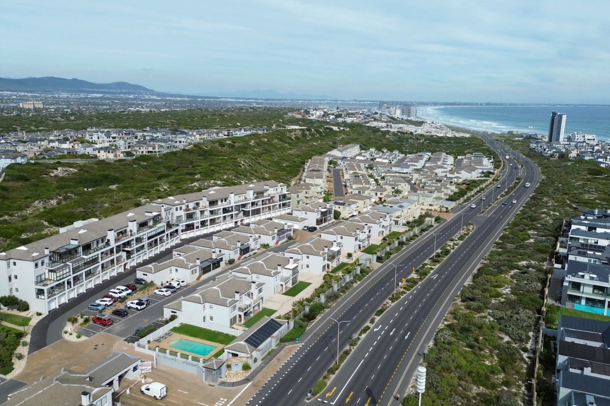 Aerial view of a coastal residential apartment complex featuring paved parking, a swimming pool, and proximity to the beach and main road.