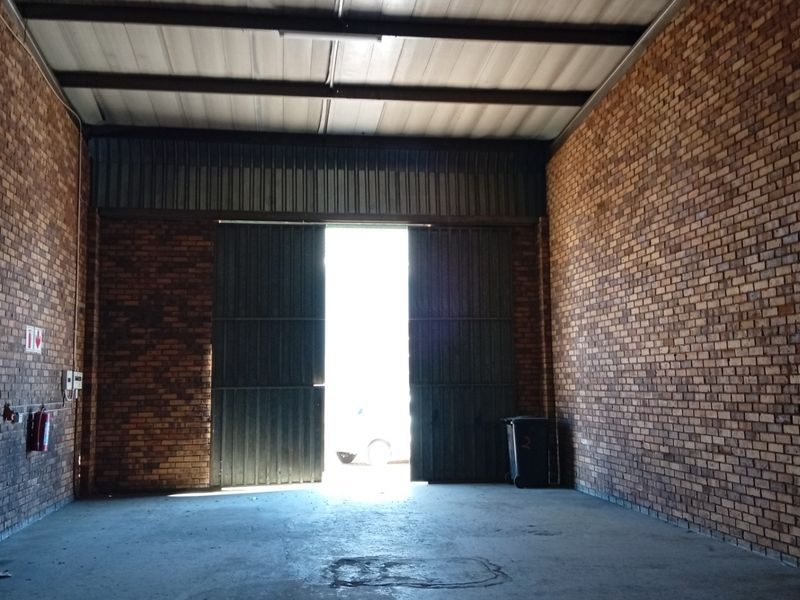 A spacious commercial garage or warehouse unit featuring exposed brick walls, concrete flooring, and high-clearance steel roof trusses with a large metal access door.