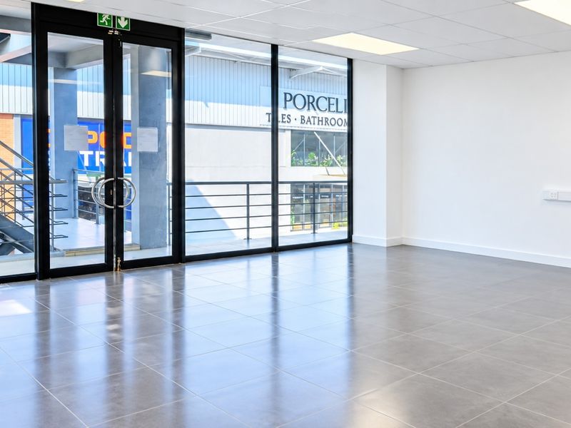A bright, modern, and empty commercial office space featuring large glass windows, tiled flooring, and a suspended ceiling with recessed lighting.