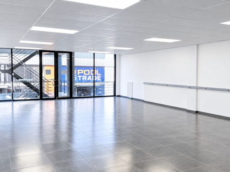 A bright, modern, and vacant commercial office space featuring tiled flooring, a suspended ceiling with recessed lighting, and large glass windows.