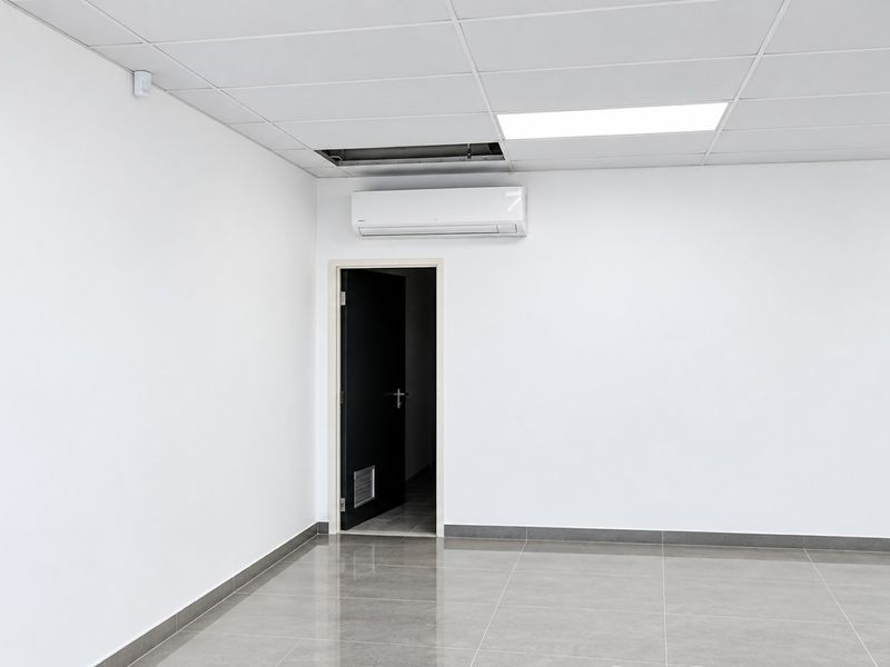 A clean, empty commercial office space featuring tiled flooring, a suspended ceiling with recessed lighting, and a wall-mounted air conditioning unit.