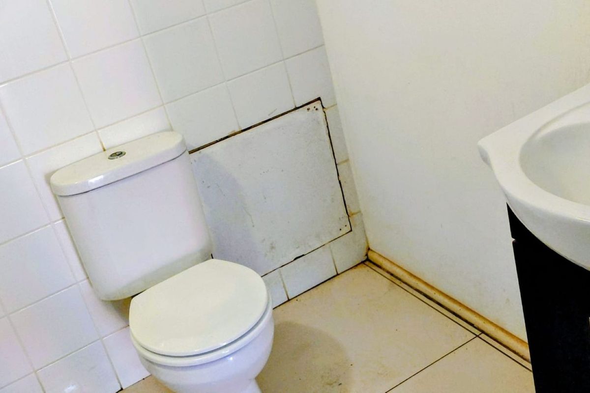 A small commercial bathroom featuring a standard toilet, vanity basin, and tiled wall section, currently showing signs of wear and staining.
