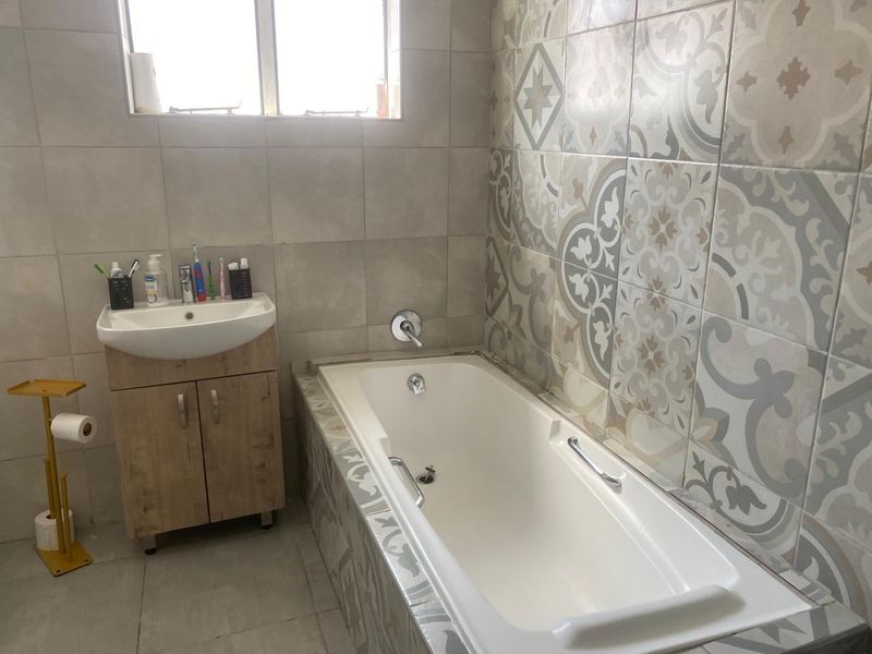 A functional bathroom featuring a built-in bathtub with decorative tiled surround, a modern vanity unit with basin, and neutral wall and floor tiling.