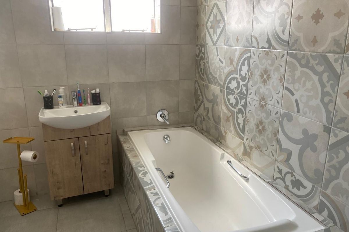 A functional bathroom featuring a built-in bathtub with decorative tiled surround, a modern vanity unit with basin, and neutral wall and floor tiling.