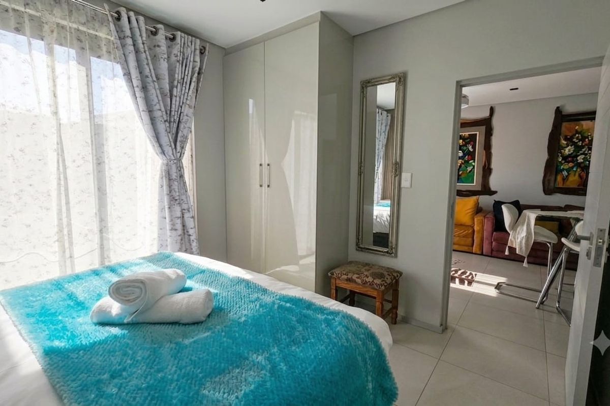 A bright bedroom featuring built-in wardrobes, tiled flooring, and a sliding glass door providing natural light, with an adjacent view into the living area.