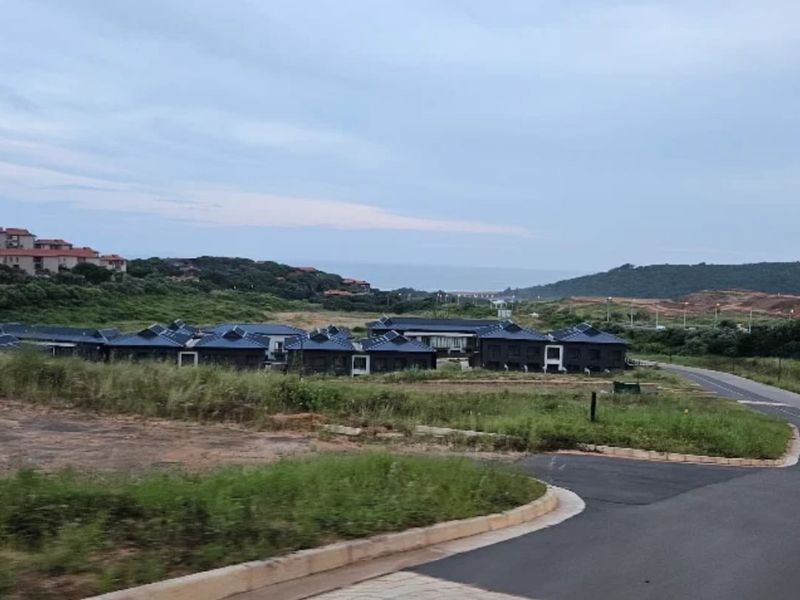 An elevated view of a residential development complex featuring multiple units with dark pitched roofs, surrounded by natural landscape and access roads.