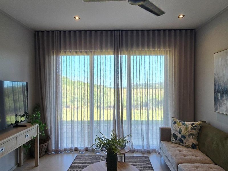 A well-lit living room featuring tiled flooring, recessed downlights, a ceiling fan, and large sliding glass doors with elegant sheer curtains.