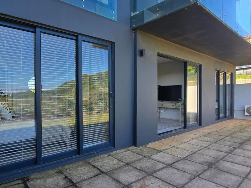 A paved ground-floor patio area featuring sliding glass doors, external air conditioning units, and a glass balustrade overlooking the garden.