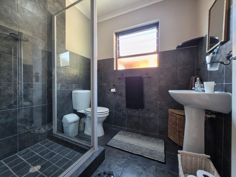A modern bathroom featuring dark grey wall and floor tiles, a glass-enclosed shower, a pedestal basin, and a louver window for ventilation.