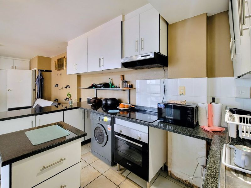 Compact kitchen featuring granite countertops, built-in oven, electric hob, extractor fan, and integrated washing machine with white cabinetry and tiled flooring.