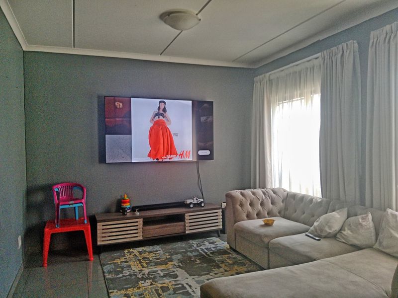 A living room featuring grey walls, tiled flooring, and a wall-mounted television, offering a functional space with natural light from the window.