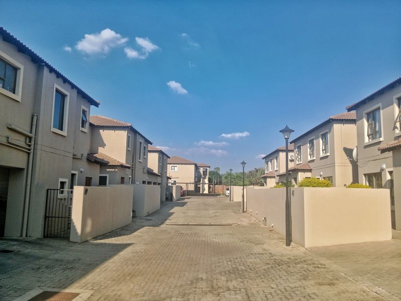 A view of a residential complex driveway featuring paved surfaces, boundary walls, and street lighting between two rows of multi-story townhouses.