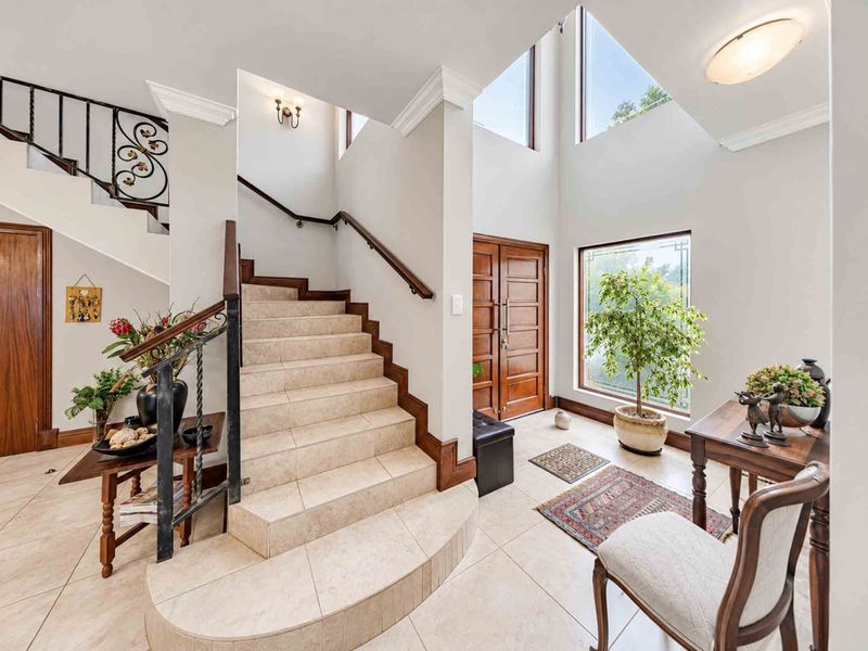 Spacious double-volume entrance hall featuring a tiled staircase with wrought iron balustrade, large feature windows, and elegant double wooden front doors.