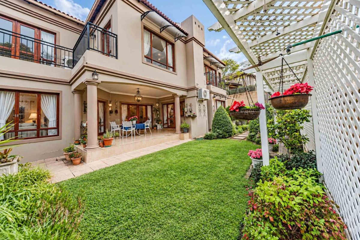 A well-maintained rear exterior view featuring a covered patio, a balcony above, a pergola, and a lush landscaped garden with a manicured lawn.