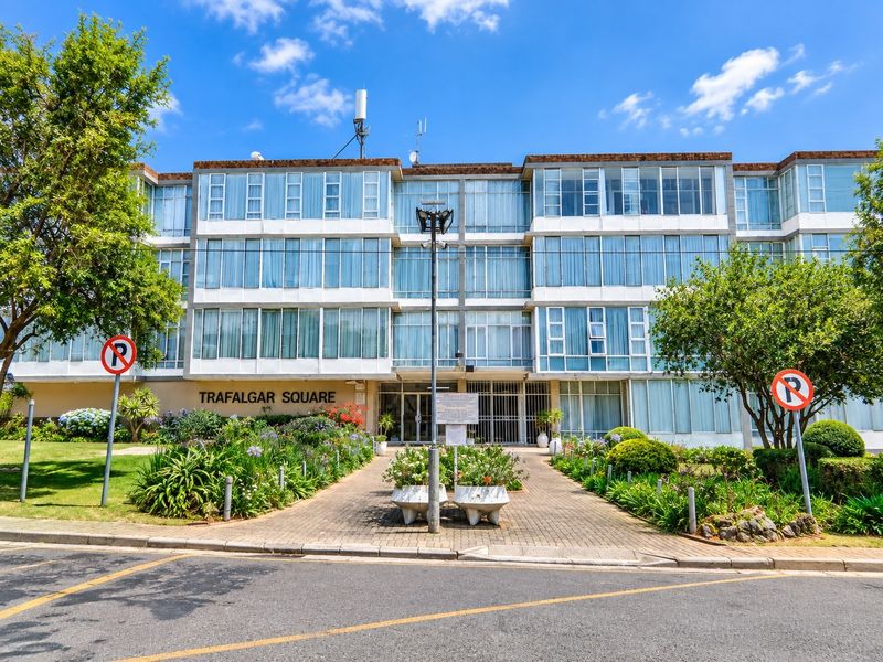 Front elevation of Trafalgar Square, a multi-story residential building featuring large glass windows, a paved entrance walkway, and established garden landscaping.