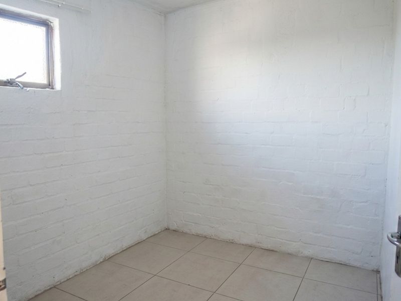 A compact, empty bedroom featuring white-painted brick walls, tiled flooring, and a small casement window, offering a blank canvas for renovation.