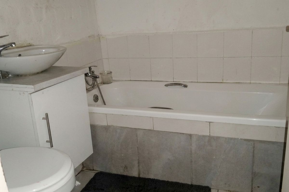 A compact bathroom featuring a built-in bathtub, vanity basin, and toilet, requiring cosmetic updates to the wall finishes and tiling.