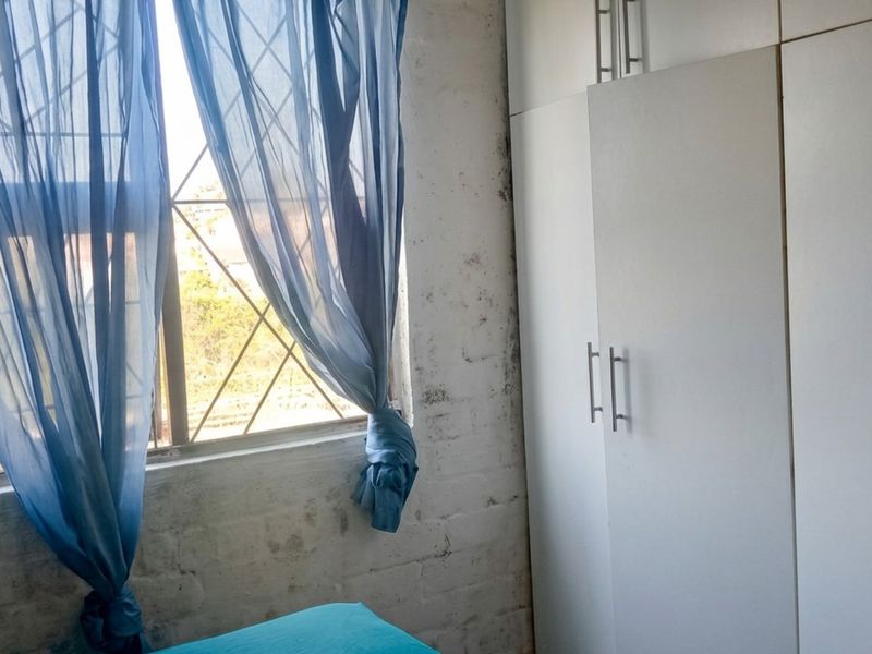 Bedroom featuring built-in cupboards and a window with security bars, showing signs of wear on the exposed brick wall finish.