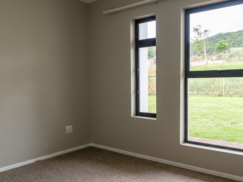 An empty bedroom featuring neutral walls, carpet flooring, and large aluminium-framed windows with views of the garden.
