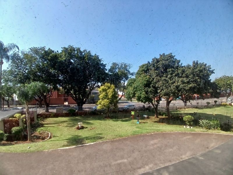 An elevated view overlooking a well-maintained garden area with mature trees, a paved driveway, and perimeter fencing.