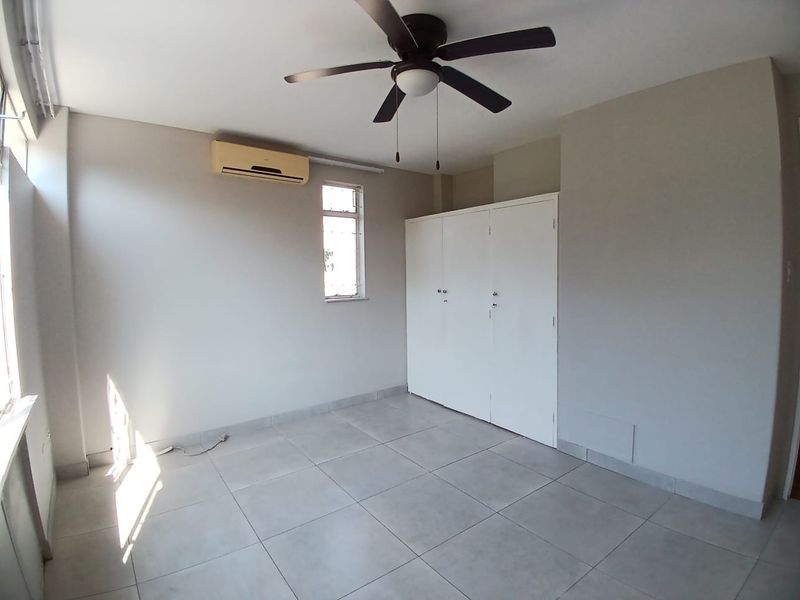A bright, empty bedroom featuring tiled flooring, a built-in wardrobe, a ceiling fan, and a wall-mounted air conditioning unit.