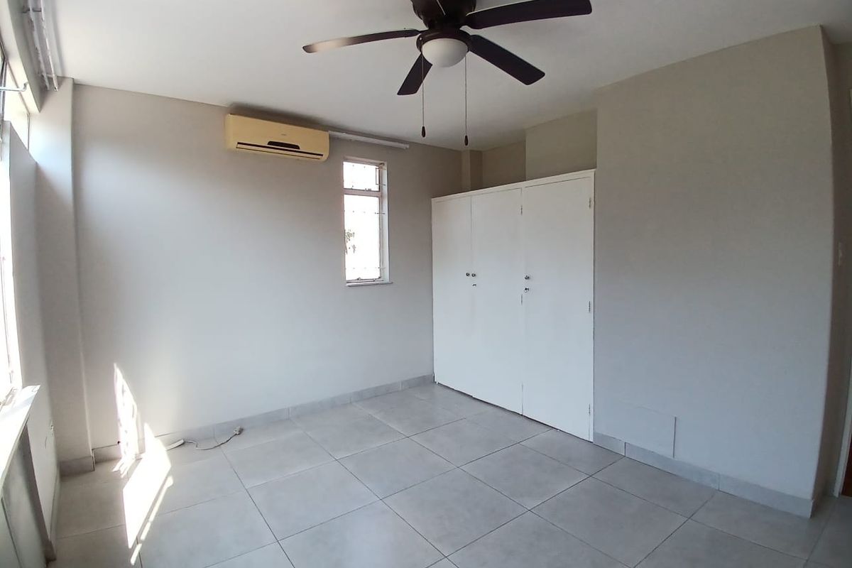 A bright, empty bedroom featuring tiled flooring, a built-in wardrobe, a ceiling fan, and a wall-mounted air conditioning unit.