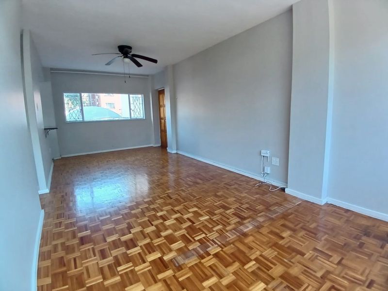 A spacious, empty living room featuring well-maintained parquet flooring, a ceiling fan, and a window with a security gate.