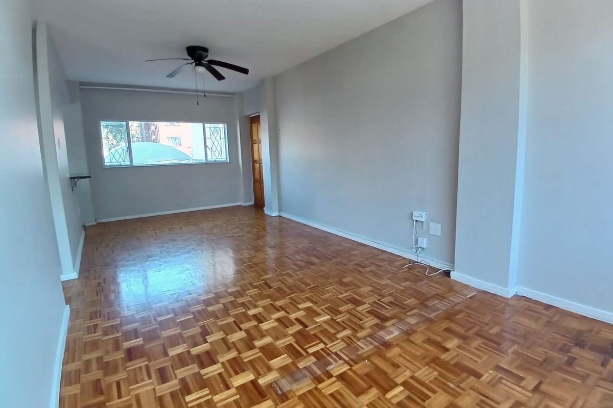 A spacious, empty living room featuring well-maintained parquet flooring, a ceiling fan, and a window with a security gate.