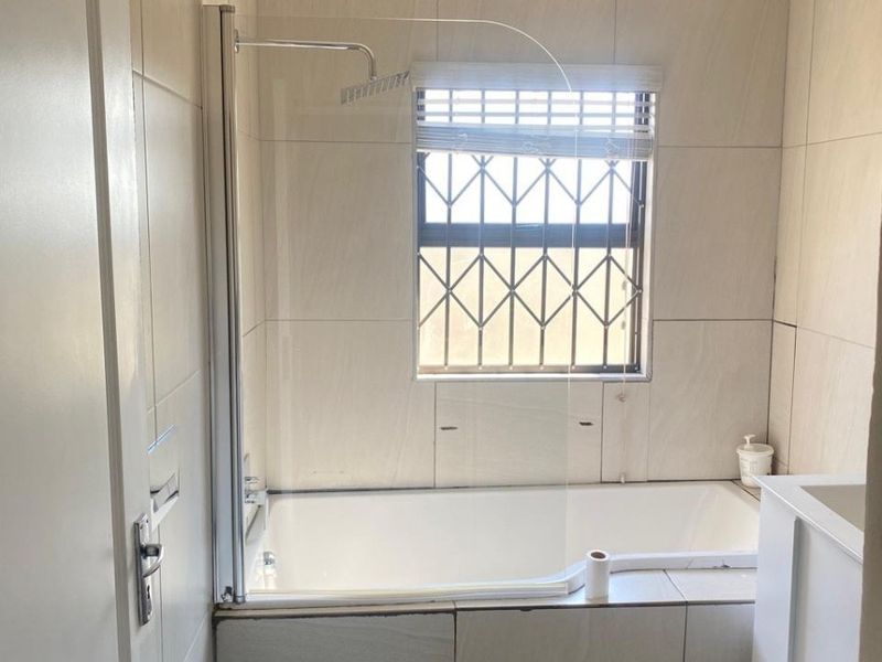 A functional bathroom featuring a bathtub with a glass shower screen, wall-mounted toilet, and vanity cabinet, finished with neutral wall tiles.