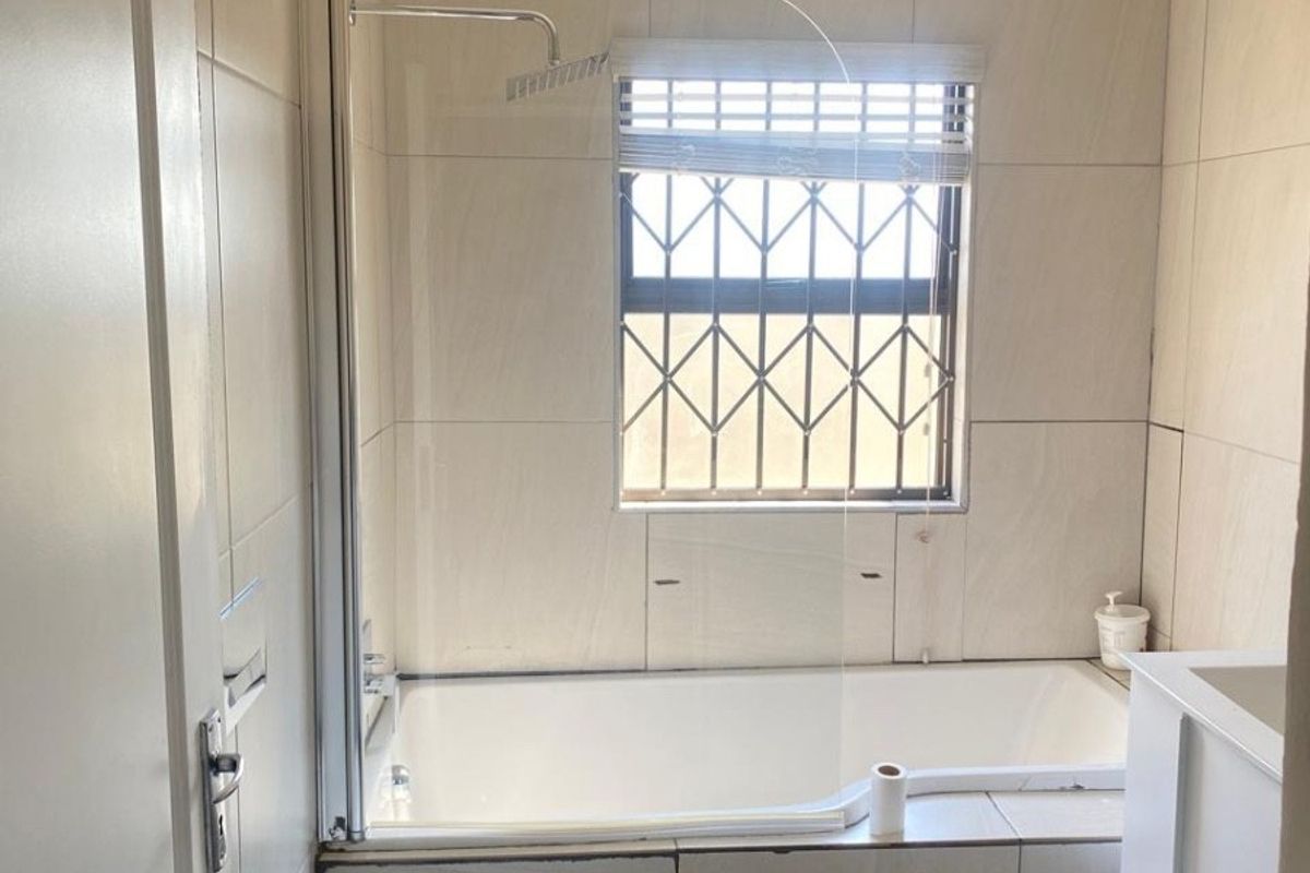 A functional bathroom featuring a bathtub with a glass shower screen, wall-mounted toilet, and vanity cabinet, finished with neutral wall tiles.