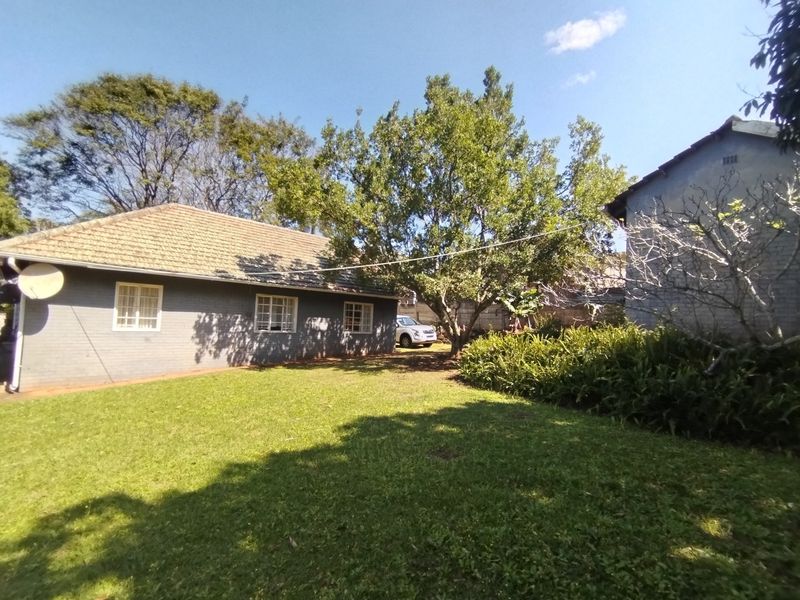 A view of the rear garden featuring a brick-walled house with a tiled roof, mature trees, and a well-maintained lawn area.