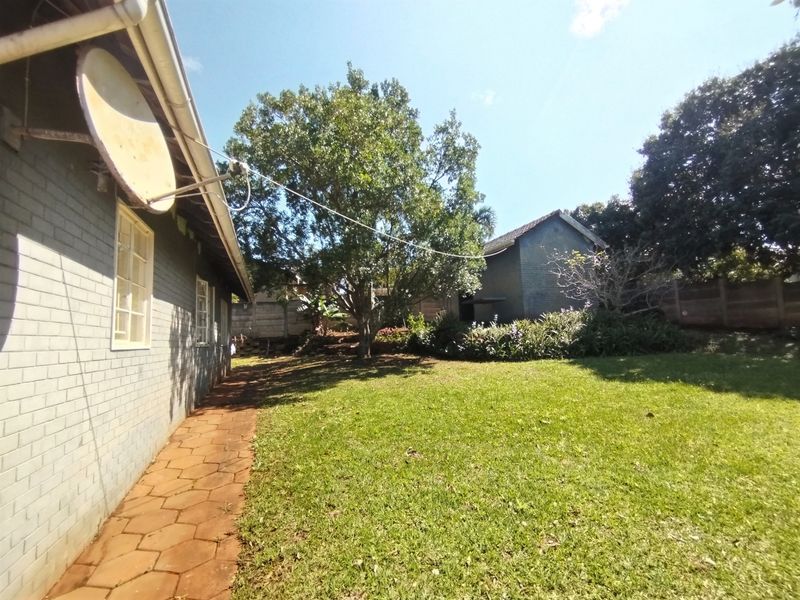 A view of the rear garden featuring a paved walkway along the brick exterior wall, mature trees, and a boundary wall.