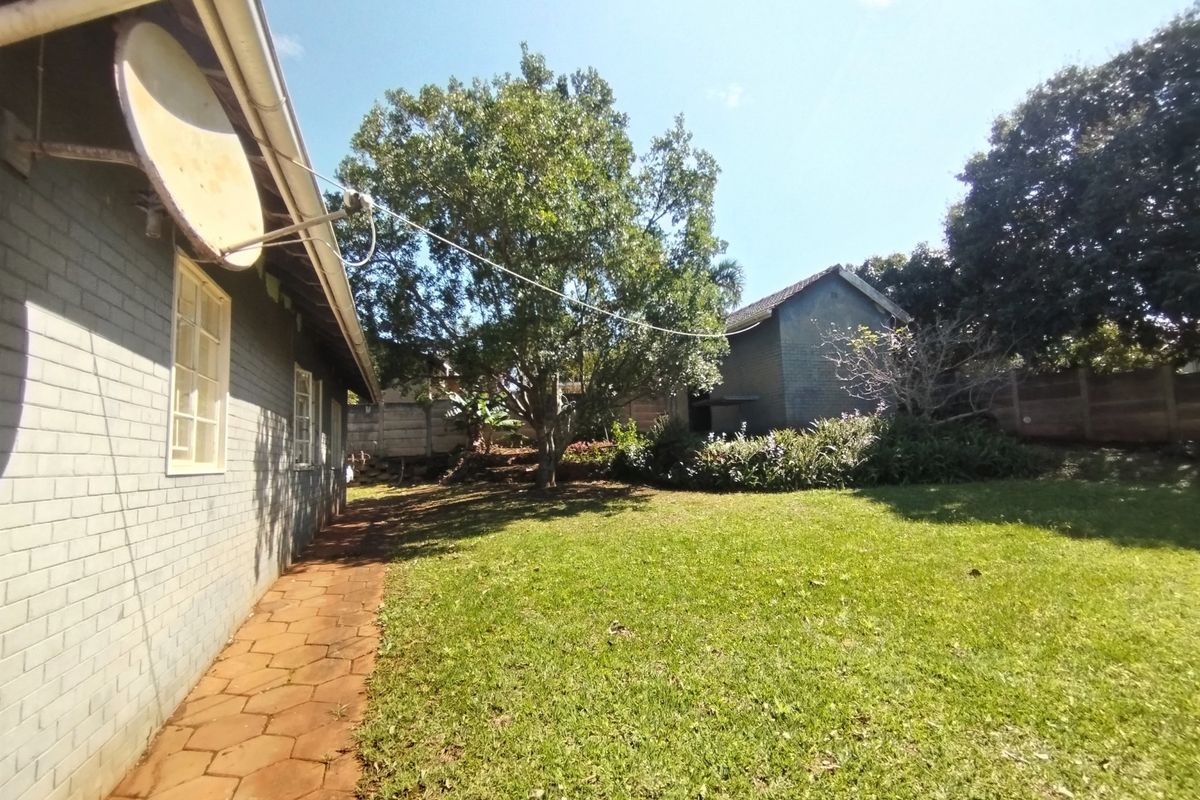 A view of the rear garden featuring a paved walkway along the brick exterior wall, mature trees, and a boundary wall.