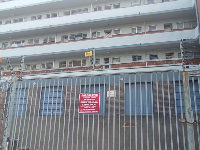 Front elevation of the Dorchester apartment complex featuring a secure gated entrance, brick facade, and multiple levels of residential units with sliding windows.
