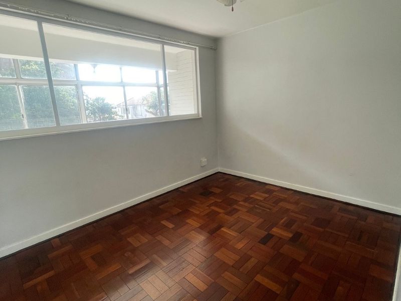 A bright, empty bedroom featuring well-maintained parquet flooring, a ceiling fan, and large sliding windows providing natural light.