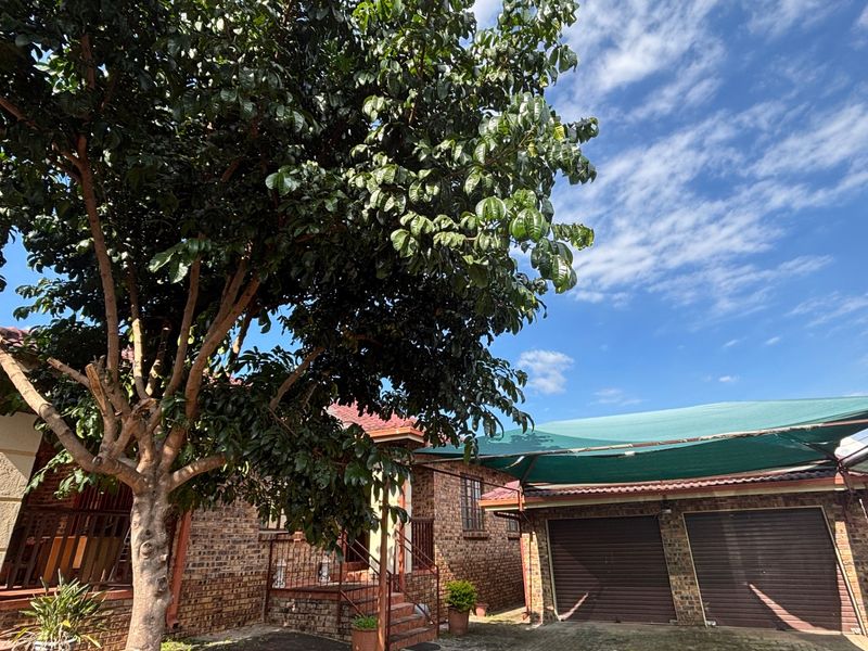 Front view of a face brick house featuring a double garage with roll-up doors, a shade cloth carport, and a mature garden tree.