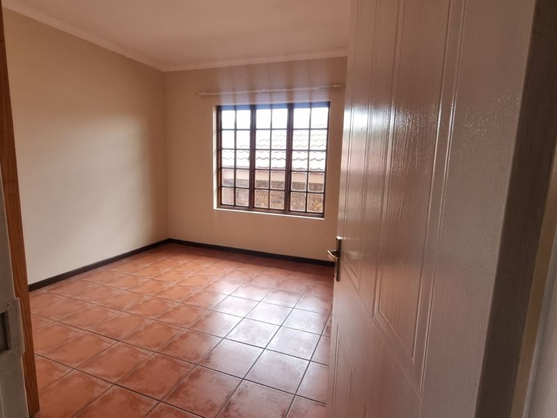 A clean, empty bedroom featuring terracotta-style floor tiles, neutral wall paint, and a window with security bars.