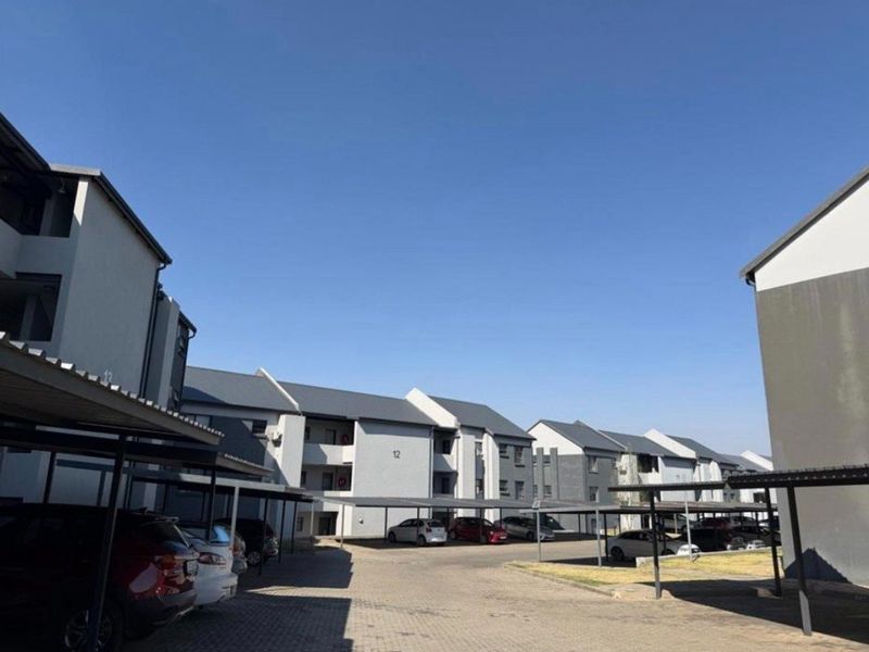 Exterior view of a residential apartment complex featuring multiple blocks, paved driveway areas, and covered parking bays under a clear blue sky.