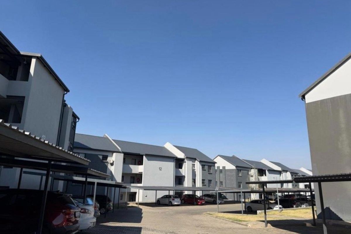 Exterior view of a residential apartment complex featuring multiple blocks, paved driveway areas, and covered parking bays under a clear blue sky.