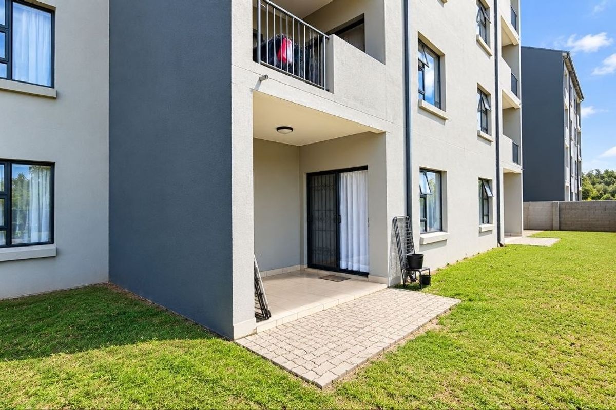 Rear view of the apartment building featuring a paved ground-floor patio, sliding door access, and a small private lawn area.